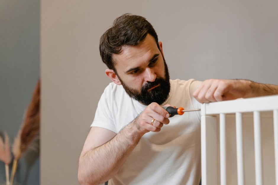 A man with a dark beard and short brown hair is carefully using a screwdriver to disassemble a white crib or small bed frame made of wood with vertical slats. He is wearing a plain white t-shirt and i
