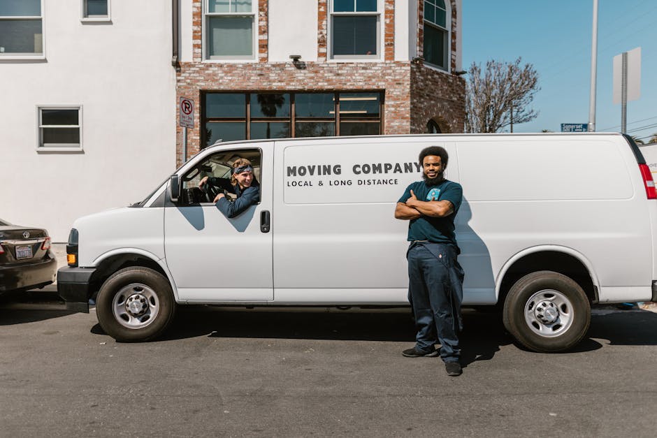 Two movers wearing uniforms and protective eyewear are loading a large piece of furniture, covered with a green moving blanket, into the back of a white van parked on a paved street. The van's rear do